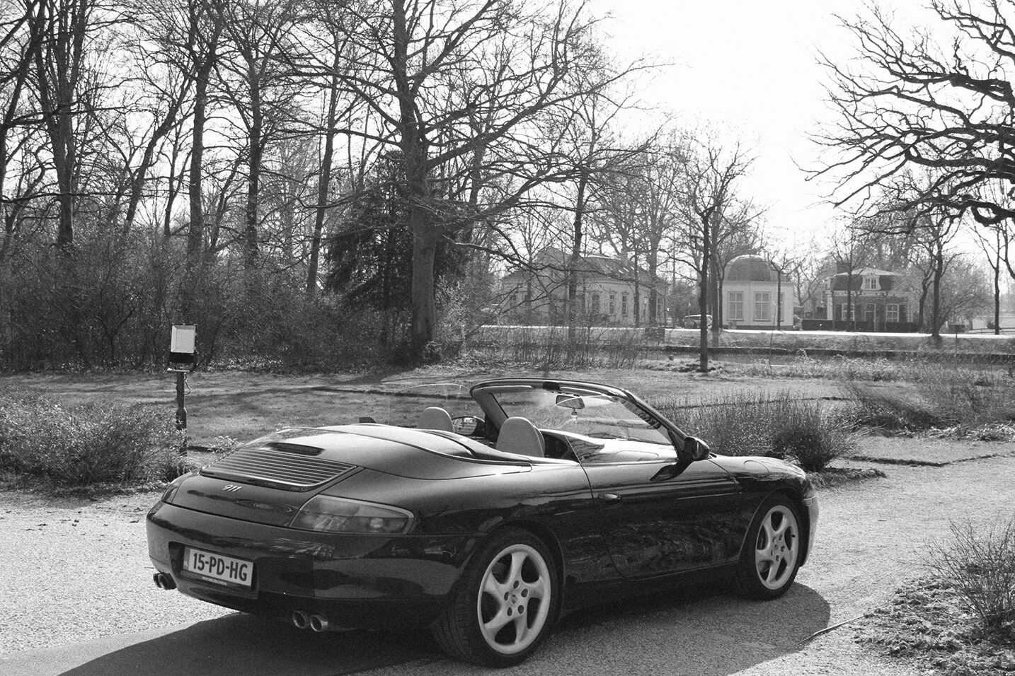 Zwart-wit foto van een Porsche 911 cabriolet geparkeerd op een landweg, omgeven door kale bomen